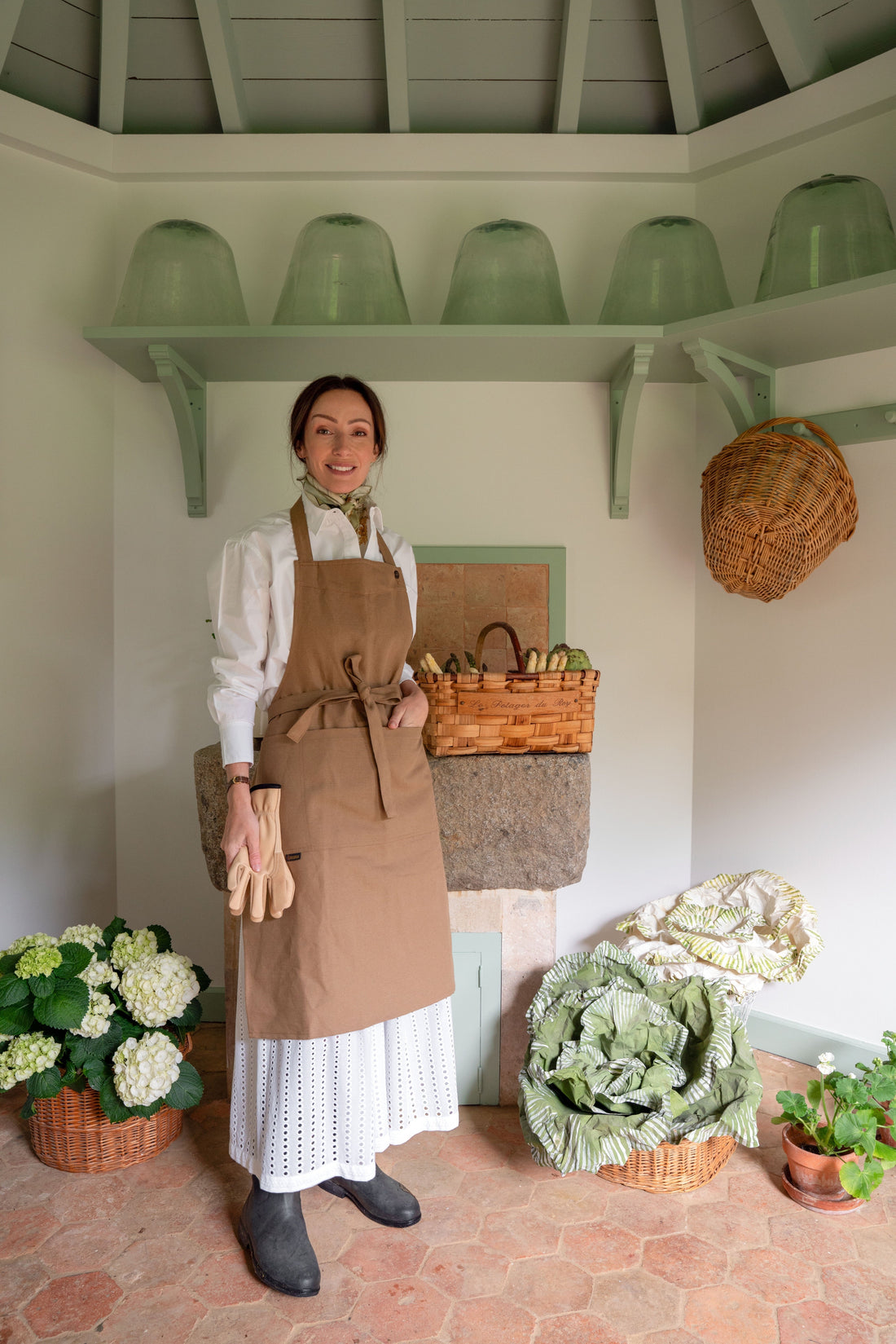 A woman wearing a white dress and a brown linen apron stands inside a rustic garden shed, surrounded by fresh white hydrangeas, large winter cabbages, and woven baskets