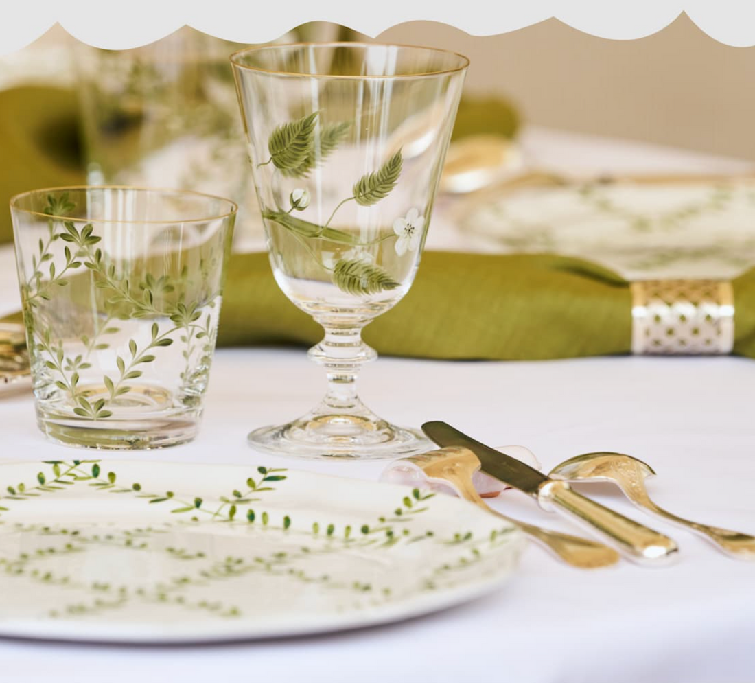 An elegant outdoor dining table set for a luncheon in a manicured French garden at Versailles, featuring green and white patterned dinnerware, fine glassware, and a lush green floral centerpiece."