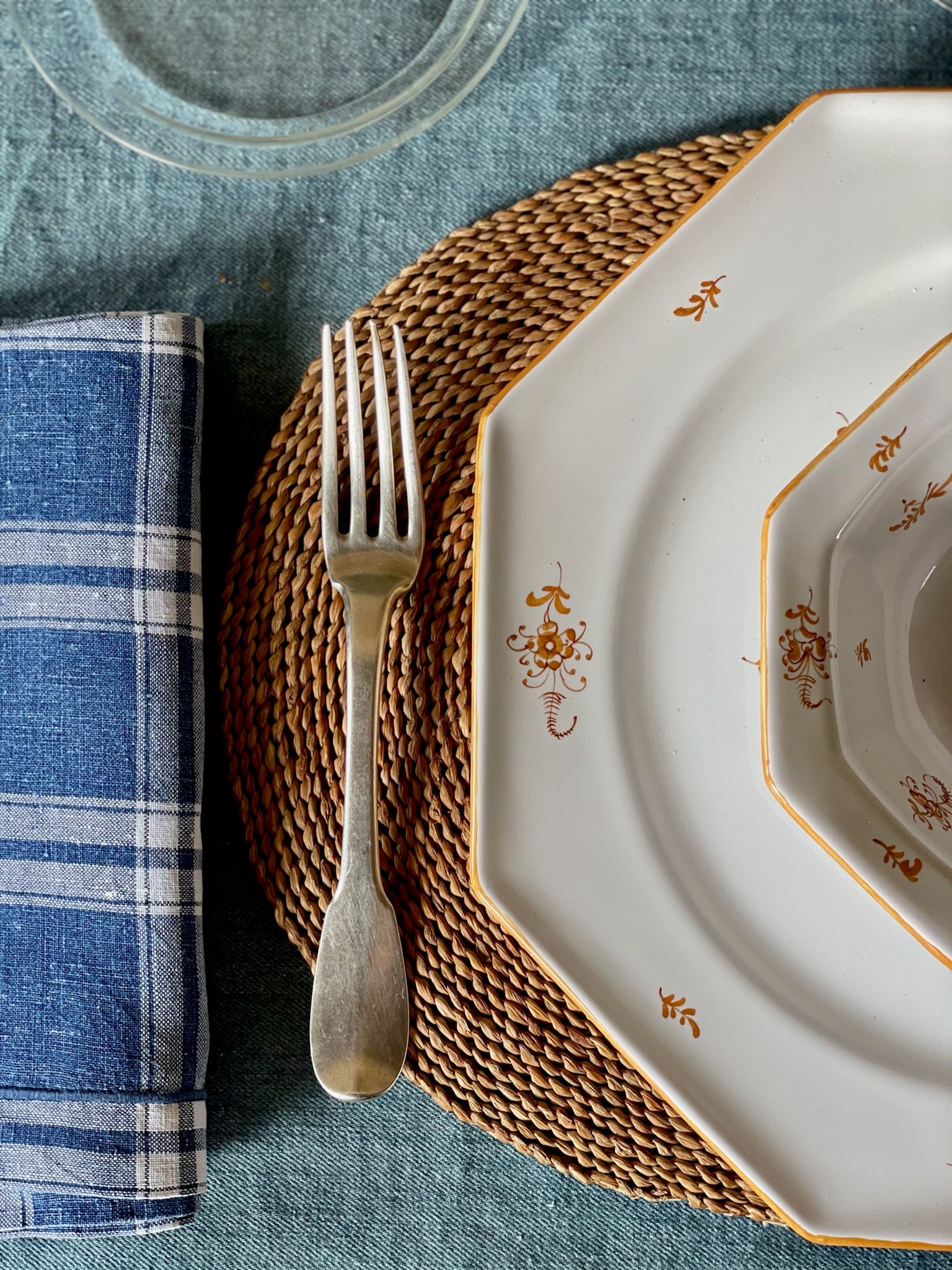 A beautifully arranged table setting featuring an octagonal white ceramic plate with a delicate ochre-brown floral border, a matching smaller bowl, and an antique silver fork, placed over a woven round placemat on a rich blue linen tablecloth.