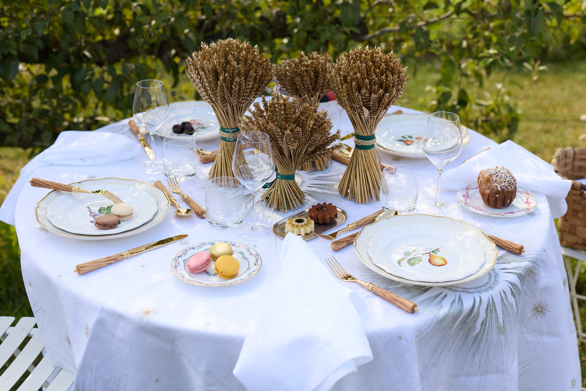An outdoor dining table styled with a white tablecloth, gold-toned cutlery, white plates with gold trim, and several small wheat bundle decorations tied with green ribbons. The setting is outdoors with green foliage in the background.