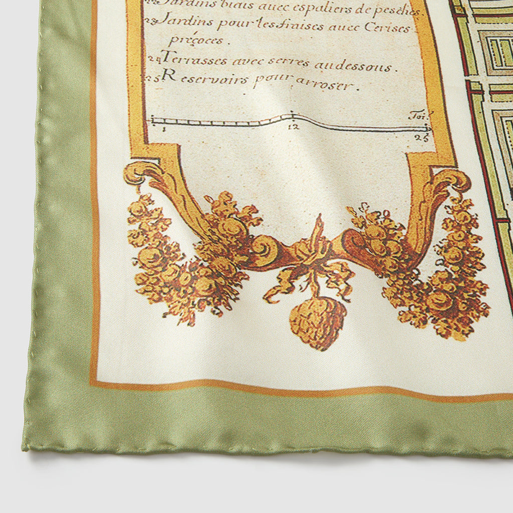 A close-up, detailed photograph of the corner of a silk scarf. It features an antique-style illustration on a cream background, showing a decorative golden-brown cartouche filled with fruit, grain, and leaves. Above the illustration, elegant French script reads "Jardins pour les fraises avec Cerises..." and "Terrasses avec serres au-dessous." The scarf is finished with a hand-rolled, solid sage green border.