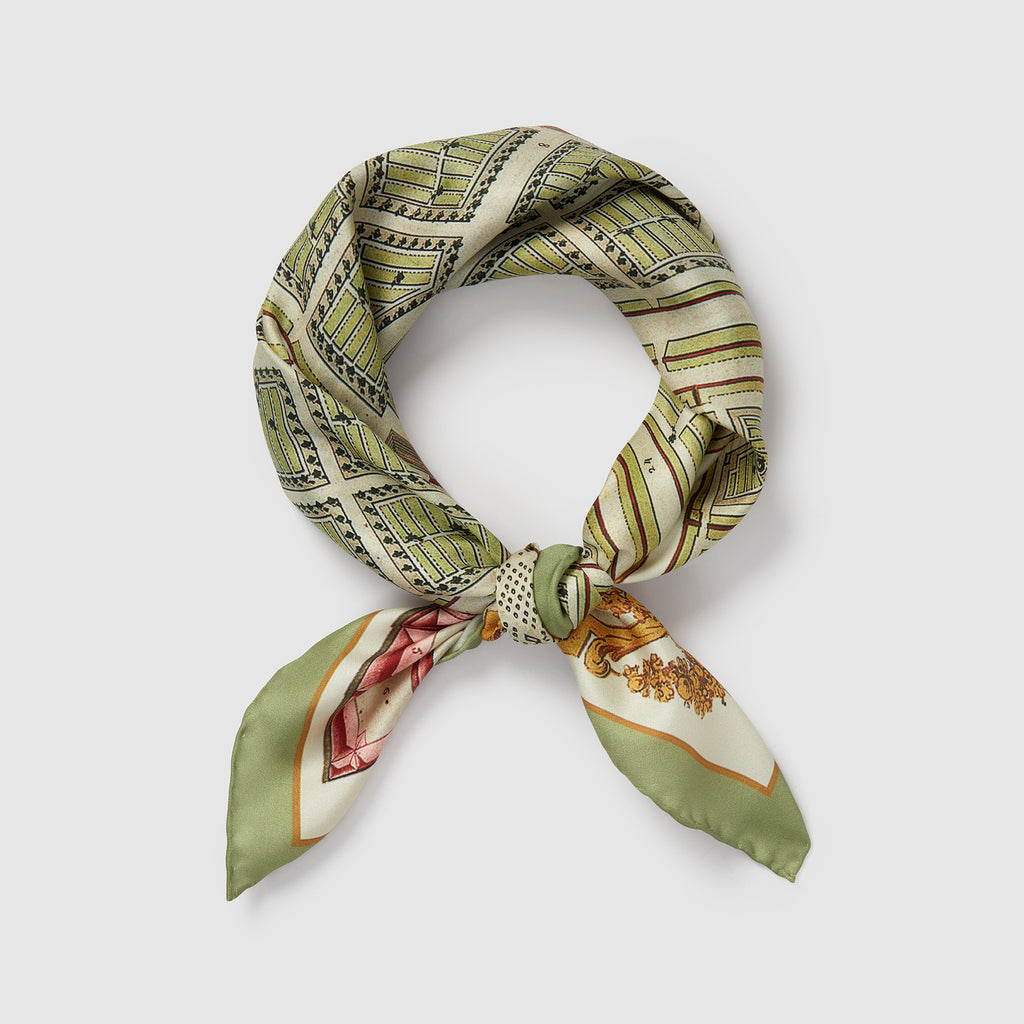 A product photograph of a square silk scarf styled into a knotted loop, showcasing its sage green borders and an illustrated map pattern. The silk fabric is draped to form a circle with the two pointed ends neatly tied together at the bottom, presented against a clean, light gray background.