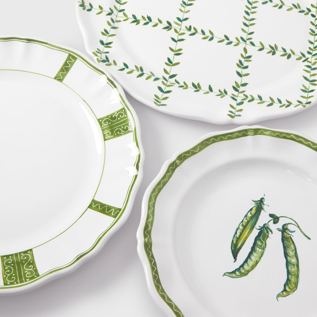A top-down product photograph showcasing three different white and green ceramic plates from the collection arranged together on a dark gray background. At the top, the charger plate features a hand-painted green leafy lattice grid design. On the left, the dinner plate displays a scalloped edge decorated with alternating green panels of swirling scrollwork and zigzag border lines. On the bottom right, the salad/dessert plate features a central illustration of three green pea pods, framed by a delicate, wavy