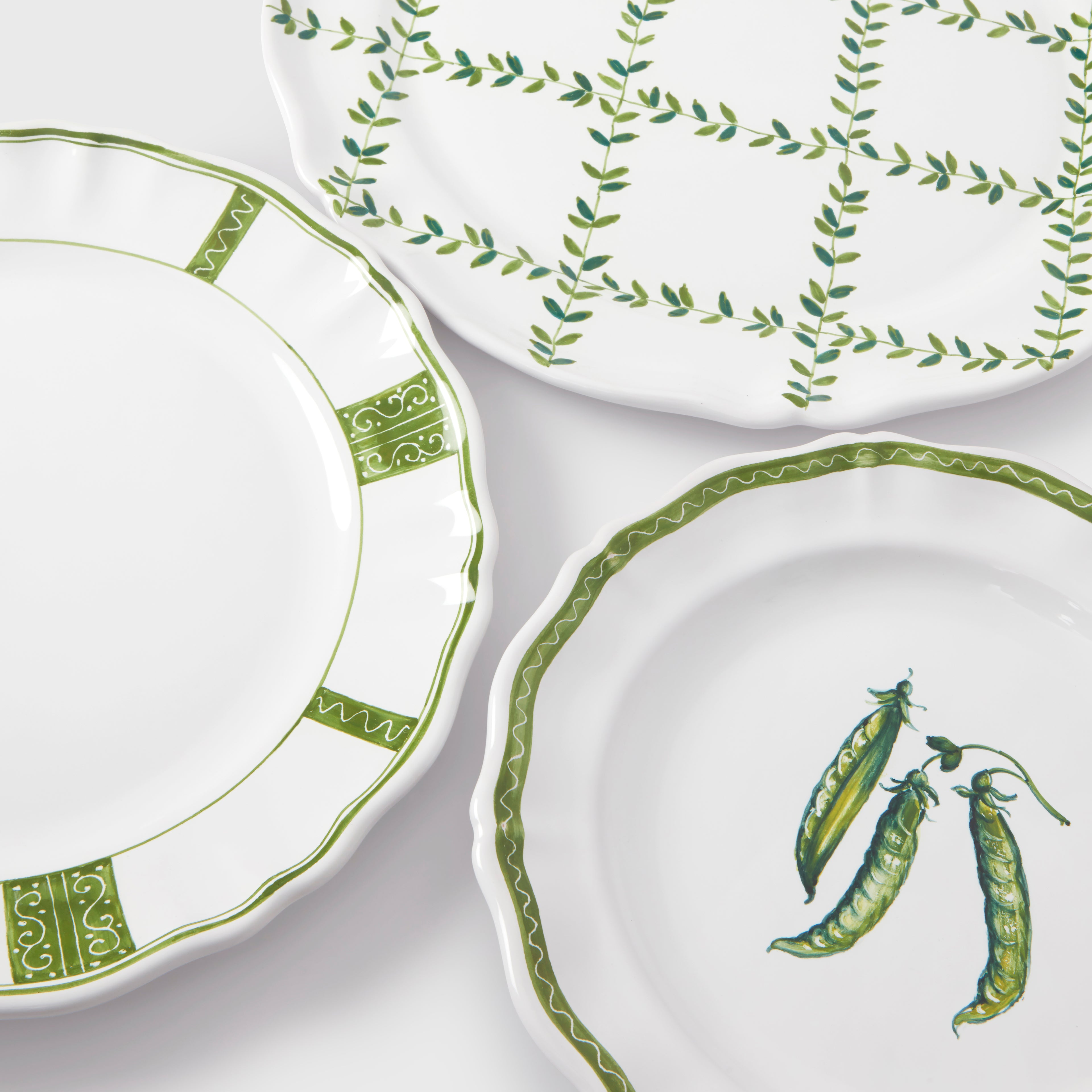 A top-down product photograph showcasing three different white and green ceramic plates from the collection arranged together on a dark gray background. At the top, the charger plate features a hand-painted green leafy lattice grid design. On the left, the dinner plate displays a scalloped edge decorated with alternating green panels of swirling scrollwork and zigzag border lines. On the bottom right, the salad/dessert plate features a central illustration of three green pea pods, framed by a delicate, wavy
