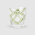 A product photograph of a clear glass tumbler with a subtle tapered shape and a gold rim, shown against a light gray background. The front of the glass is delicately hand-painted with a botanical design featuring slender, arching green branches and delicate leaves arranged in an elegant, symmetrical wreath formation.