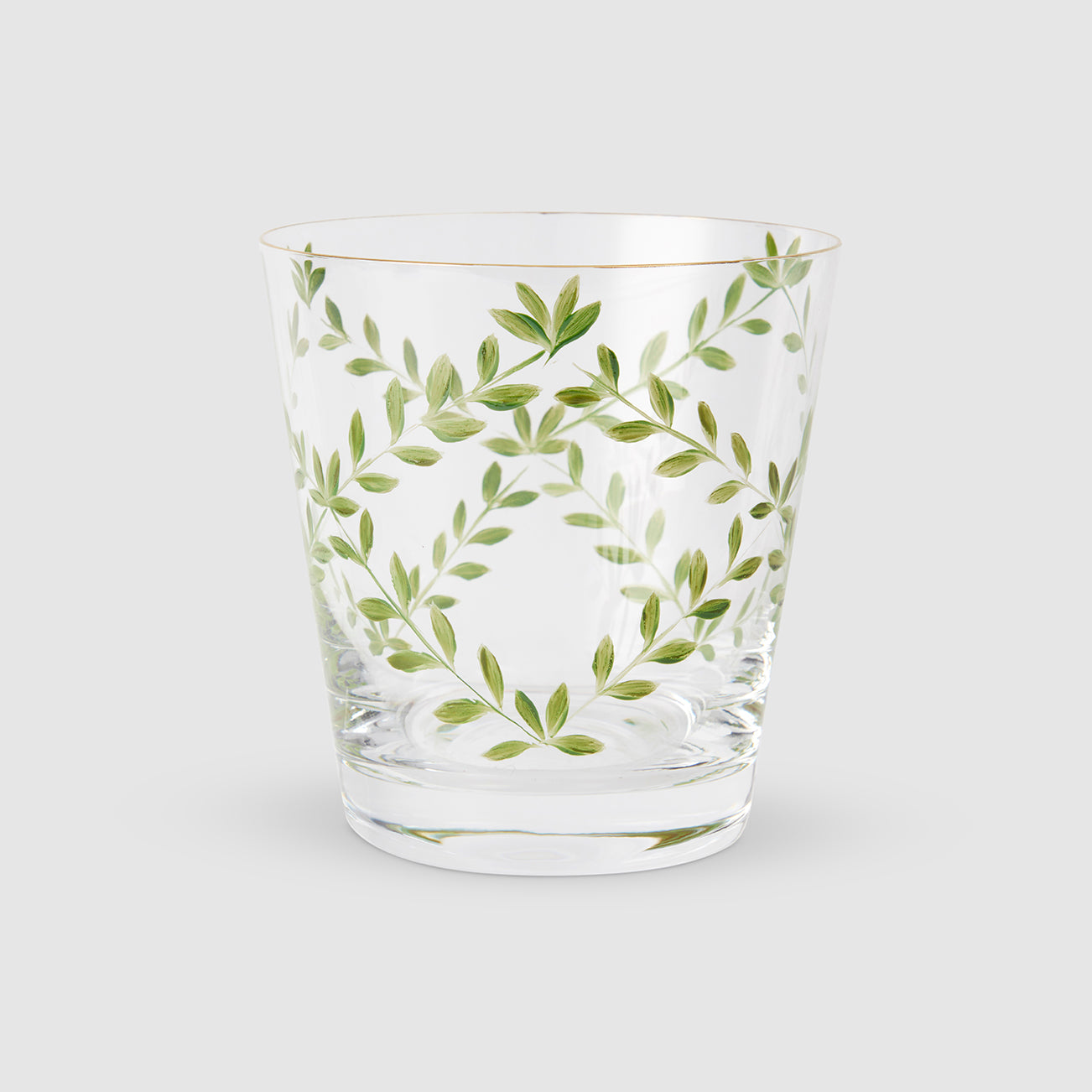 A product photograph of a clear glass tumbler with a subtle tapered shape and a gold rim, shown against a light gray background. The front of the glass is delicately hand-painted with a botanical design featuring slender, arching green branches and delicate leaves arranged in an elegant, symmetrical wreath formation.