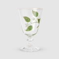 A product photograph of an elegant, clear glass wine or water glass with a footed stem and a fine gold rim, set against a clean, light gray background. The front of the glass is delicately hand-painted with a botanical design featuring a slender green branch with leaves and small white blossoms.