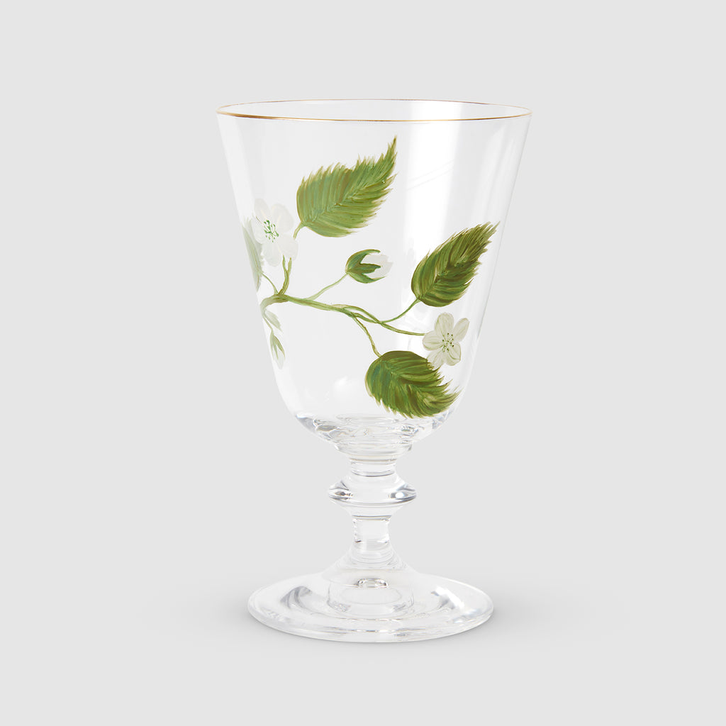 A product photograph of an elegant, clear glass wine or water glass with a footed stem and a fine gold rim, set against a clean, light gray background. The front of the glass is delicately hand-painted with a botanical design featuring a slender green branch with leaves and small white blossoms.