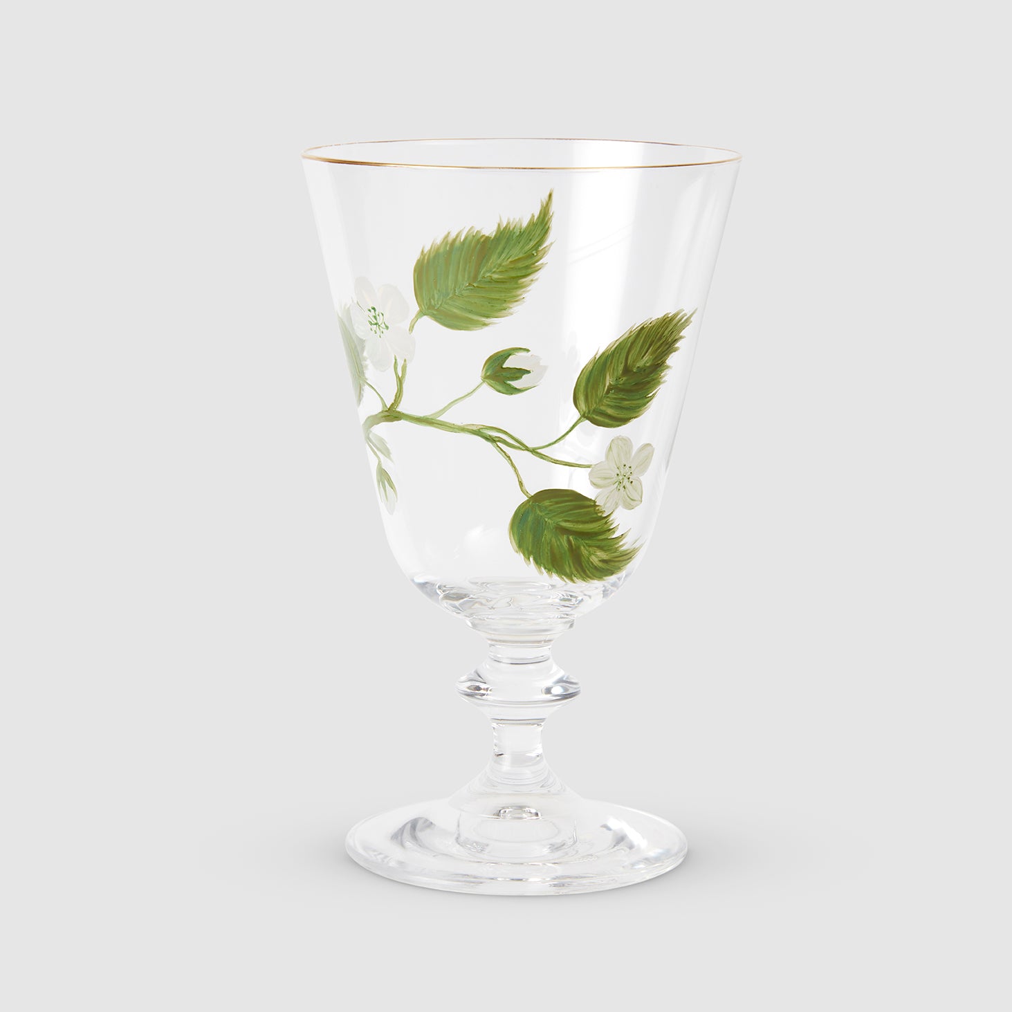 A product photograph of an elegant, clear glass wine or water glass with a footed stem and a fine gold rim, set against a clean, light gray background. The front of the glass is delicately hand-painted with a botanical design featuring a slender green branch with leaves and small white blossoms.