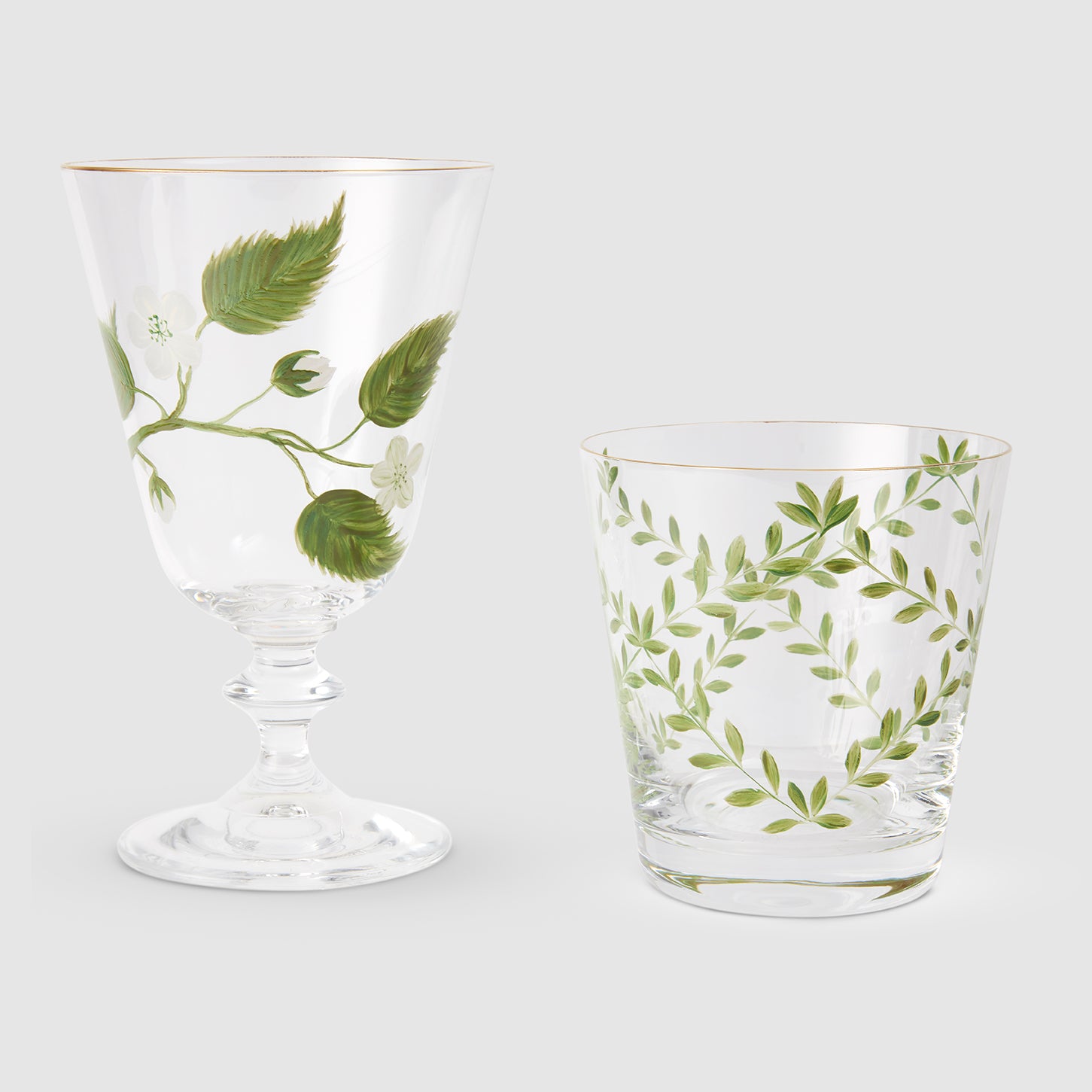 A product photograph displaying two clear glassware pieces side by side against a light gray background, both featuring gold rims and delicate hand-painted botanical designs. On the left is an elegant, stemmed footed glass decorated with a branch of green leaves, white blossoms, and a single green apple. On the right is a matching tapered tumbler adorned with a symmetrical wreath-like pattern of slender green branches and fine leaves.