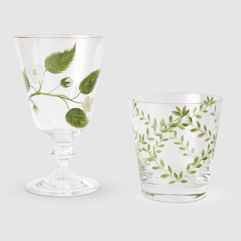product photograph displaying two clear glassware pieces side by side against a light gray background, both featuring gold rims and delicate hand-painted botanical designs. On the left is an elegant, stemmed footed glass decorated with a slender branch of green leaves and small white blossoms. On the right is a matching tapered tumbler adorned with a symmetrical wreath-like pattern of slender green branches and fine leaves.