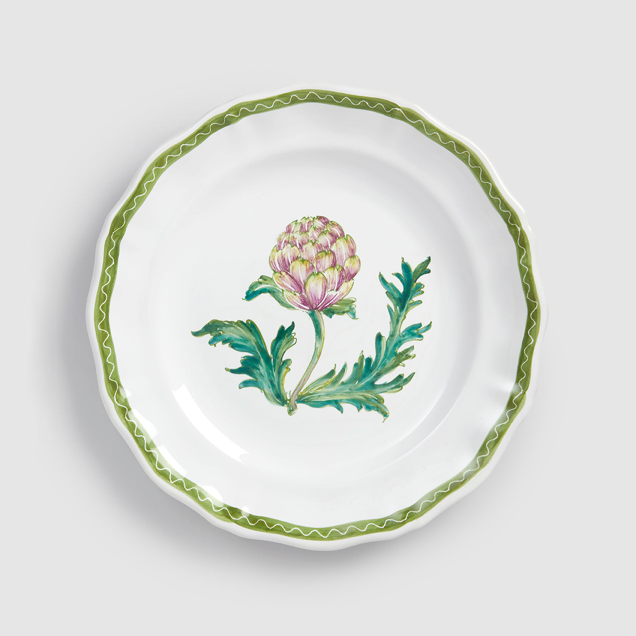 A top-down product photograph of a white round ceramic salad or dessert plate with a delicately scalloped rim, shown against a light gray background. The center features a hand-painted botanical illustration of an artichoke head in shades of green and purple-pink, surrounded by slender green leaves. A thin, wavy green zigzag line traces the exact contour of the scalloped edge.