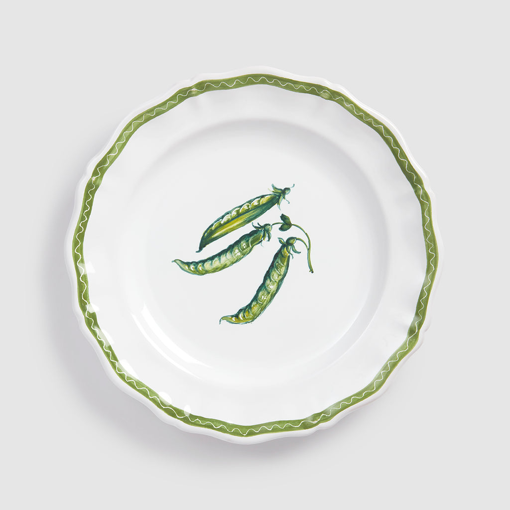 A top-down product photograph of a white round ceramic salad or dessert plate with a delicately scalloped rim, shown against a light gray background. The center features a hand-painted botanical illustration of three green pea pods clustered together, with one pod partially open to show the small peas inside. A thin, wavy green zigzag line traces the exact contour of the scalloped edge.