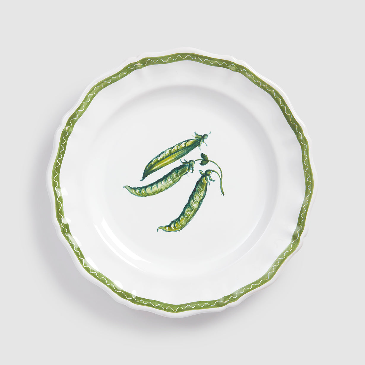 A top-down product photograph of a white round ceramic salad or dessert plate with a delicately scalloped rim, shown against a light gray background. The center features a hand-painted botanical illustration of three green pea pods clustered together, with one pod partially open to show the small peas inside. A thin, wavy green zigzag line traces the exact contour of the scalloped edge.