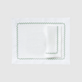 A flat lay photograph of the Celadon Placemat and Napkin Set in white on a dark, grid-patterned background. The set features a rectangular white linen placemat laid flat with a matching white linen napkin folded neatly on top. Both pieces are decorated with a delicate, embroidered green wavy scalloped border along their edges.