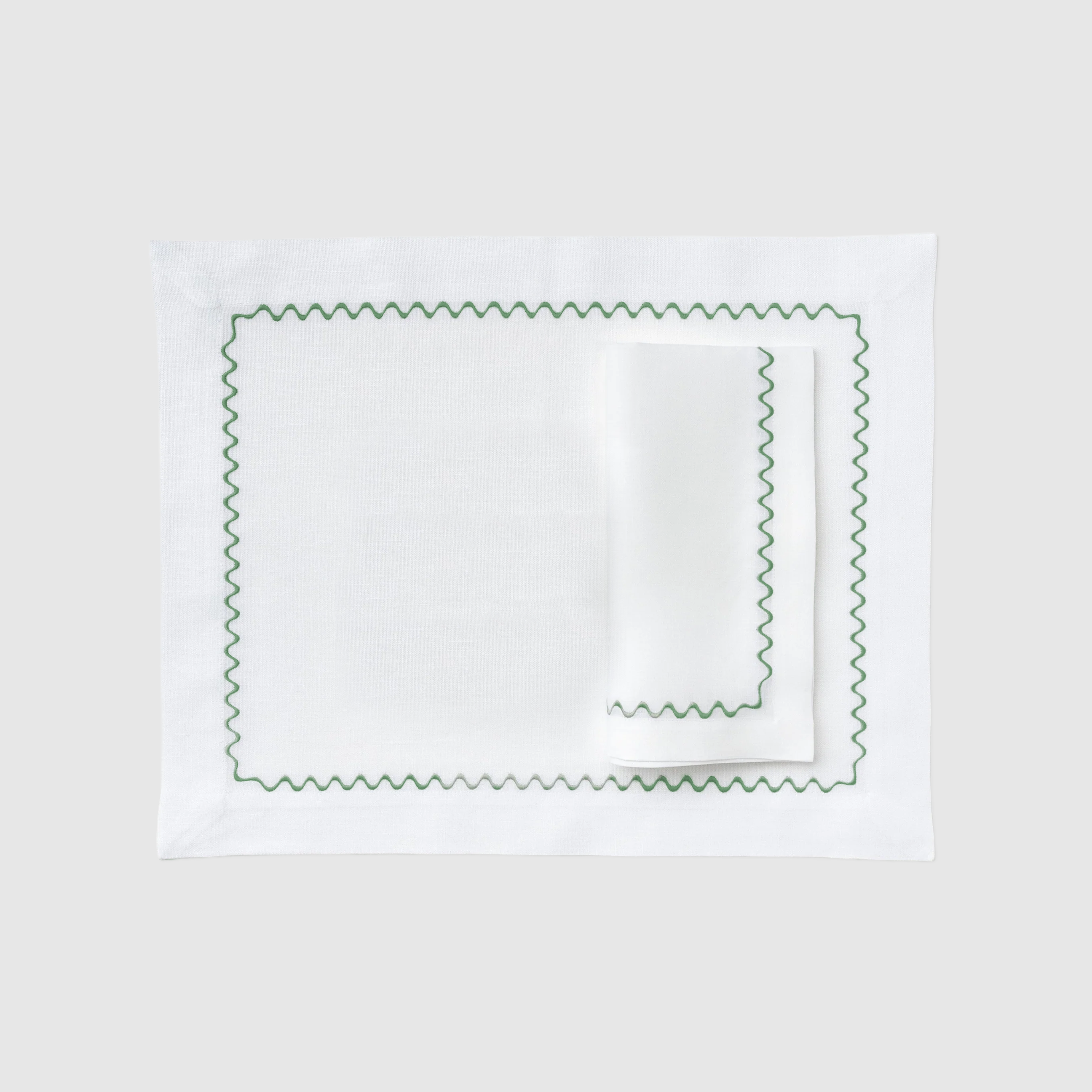 A flat lay photograph of the Celadon Placemat and Napkin Set in white on a dark, grid-patterned background. The set features a rectangular white linen placemat laid flat with a matching white linen napkin folded neatly on top. Both pieces are decorated with a delicate, embroidered green wavy scalloped border along their edges.