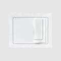 A flat lay photograph of the Celadon Placemat and Napkin Set in white on a dark, grid-patterned background. The set features a rectangular white linen placemat laid flat with a matching white linen napkin folded neatly on top. Both pieces are decorated with a delicate, embroidered green wavy scalloped border along their edges.