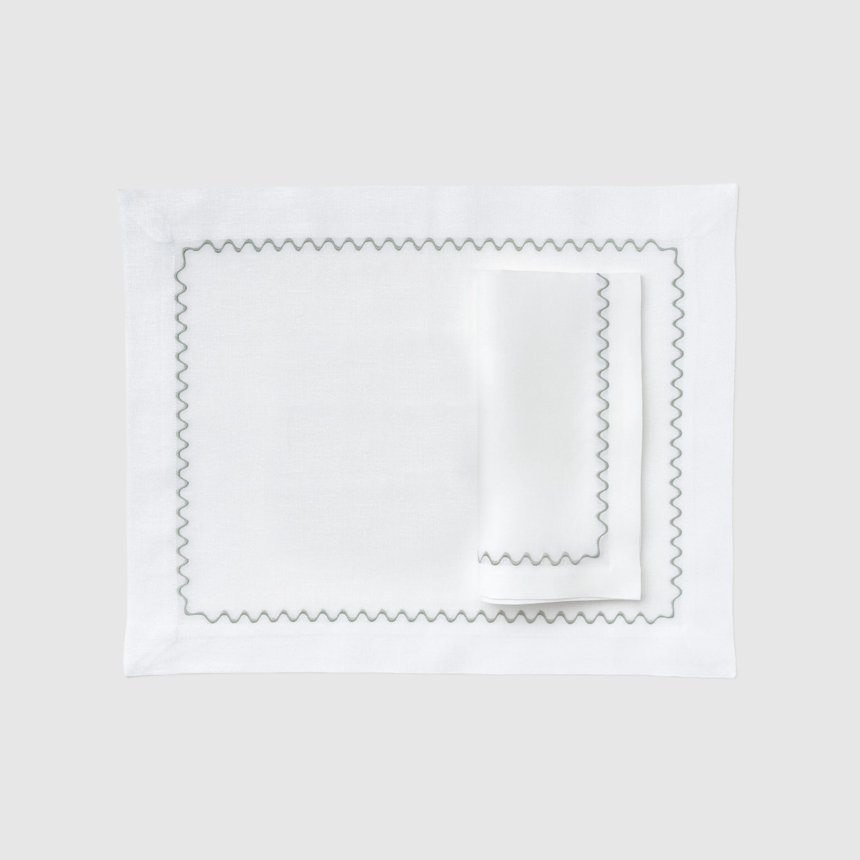 A flat lay photograph of the Celadon Placemat and Napkin Set in white on a dark, grid-patterned background. The set features a rectangular white linen placemat laid flat with a matching white linen napkin folded neatly on top. Both pieces are decorated with a delicate, embroidered green wavy scalloped border along their edges.
