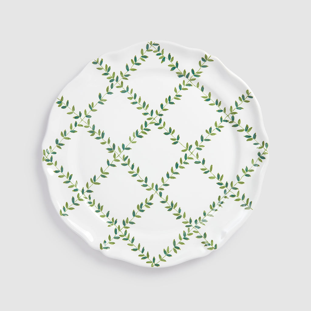 A product photograph of a white round ceramic charger plate, shown flat from a top-down view against a clean, light gray background. The surface of the plate features a hand-painted botanical design with green leafy vines intersecting to form a lattice or diamond grid pattern across the center.