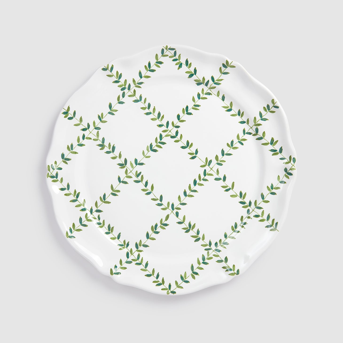 A product photograph of a white round ceramic charger plate, shown flat from a top-down view against a clean, light gray background. The surface of the plate features a hand-painted botanical design with green leafy vines intersecting to form a lattice or diamond grid pattern across the center.
